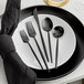 A five-piece set of black brushed stainless steel flatware, including two forks, a knife, and two spoons, displayed on a white plate with a black napkin.