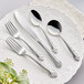 A five-piece set of stainless steel flatware with ornate handles, including two forks, a knife, and two spoons, displayed on a white decorative plate.