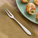 An extra heavy weight stainless steel escargot fork with two tines, placed on a napkin next to a plate of escargot.