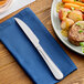 A stainless steel steak knife with a serrated edge resting on a blue napkin next to a plate of food.