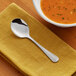A stainless steel bouillon spoon placed on a yellow napkin next to a bowl of soup.