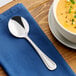 A stainless steel bouillon spoon with a decorative handle, placed on a blue napkin next to a bowl of soup.