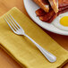 A stainless steel European table fork placed on a yellow napkin next to a plate of breakfast food.