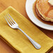 A stainless steel dinner fork placed on a yellow napkin next to a plate of pancakes with syrup and butter.