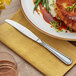 A stainless steel heavy weight dinner knife with a simple, elegant handle design placed on a yellow napkin next to a plate of food.