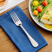 A stainless steel dinner fork placed on a blue napkin next to a plate of food.