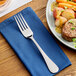 A stainless steel European dinner fork placed on a blue napkin next to a plate of food.