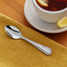 A stainless steel demitasse spoon with a simple, elegant design placed on a yellow napkin next to a cup of tea with lemon.