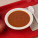 A tan Kingline melamine grapefruit bowl filled with soup and a spoon on a red tray with two apples.