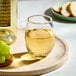 A clear, stemless wine glass filled with white wine, placed on a round tray next to a bottle and some grapes.