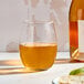 A clear, stemless wine glass filled with a golden liquid, placed on a light surface next to a wine bottle.