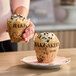A tulip-shaped baking cup with a newspaper print design, holding a cupcake topped with white frosting and chocolate chips.