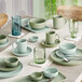 A dining table is set with various pastel-colored ceramic dishes, bowls, mugs, and glassware in shades of green and blue.
