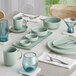 A neatly set dining table featuring light green ceramic dishes, bowls, mugs, and blue-tinted glassware.