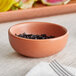 A small, round, terra cotta-colored bowl with a matte finish containing a dark, crumbly substance, placed on a light fabric surface.