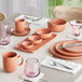 A set dining table featuring terra cotta-colored dishware, including bowls, plates, mugs, and pink-tinted glassware.