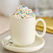 A white mug filled with whipped cream and colorful sprinkles, placed on a matching saucer with a spoon.