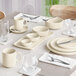 A dining table set with cream-colored porcelain dishes, bowls, mugs, and various glassware, arranged neatly with silverware and white napkins.