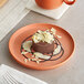 A small chocolate dessert topped with white chocolate shavings is served on an orange matte plate with decorative chocolate and cream sauces.