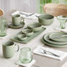 A dining table set with sage green matte finish porcelain dinnerware, including bowls, plates, mugs, and green-tinted glassware.