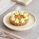 A small tart topped with whipped cream, lemon slices, and mint leaves is served on a round, matte white plate.