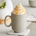 A gray ceramic mug filled with a beverage topped with whipped cream and caramel drizzle, placed on a white coaster.
