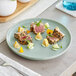 A light blue matte plate with a gourmet arrangement of seared tuna slices, lemon wedges, cucumber pieces, green sauce dollops, and edible flowers.
