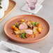 A terracotta-colored porcelain plate with a gourmet dish featuring sliced meat, roasted vegetables, microgreens, and sauce dots, artfully arranged.