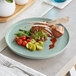 A blue matte porcelain plate with a gourmet meal featuring lamb chops, potatoes, cherry tomatoes, asparagus, microgreens, and sauce.