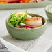 A small green matte-finish bowl containing a seared scallop, greens, radish slices, and a lemon wedge.