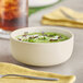 A matte white bowl filled with green soup and garnished with microgreens, placed on a table with a yellow napkin and a spoon nearby.