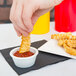 A hand dipping a french fry into a white Carlisle fluted plastic ramekin filled with ketchup.