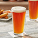 A clear 20 oz. rim tempered mixing glass filled with amber beer, placed on a white napkin on a marble surface.