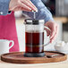 A person is pressing down the plunger of a glass and metal French press filled with coffee, placed on a wooden tray with a spoon nearby.