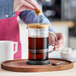 A person is pressing down the plunger of a glass and wood French press filled with coffee, placed on a wooden tray with a spoon nearby.