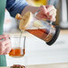 A person is pouring tea or coffee from a glass French press with a wooden lid into a clear glass cup.