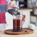 A person is pressing down the plunger of a glass French press filled with coffee on a wooden tray, with a cup, spoon, and bowl of sugar nearby.