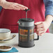 A person is pressing down the plunger of a black French press filled with tea or coffee on a light-colored table, with a bowl, spoon, and cup nearby.