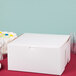 A plain white cardboard bakery box with a closed lid, measuring 12 by 12 by 6 inches.