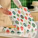 A person is opening a festive bakery box decorated with colorful ornament patterns on a kitchen counter.