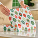 A person is closing a festive bakery box decorated with colorful holiday ornament patterns on a kitchen counter.
