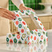 A person is closing a festive bakery box with a clear window and colorful ornament patterns on a kitchen counter.