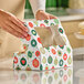 A person is closing a festive bakery box with a holiday ornament pattern and a clear window on top, placed on a kitchen counter.