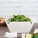 A white square porcelain bowl with handles filled with salad and vegetables on a table.