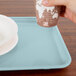 A hand holding a cup over a rectangular sky blue Cambro tray with a plate and bowl on it.