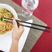 A hand holding a pair of black Town plastic chopsticks over a bowl of noodles.