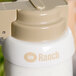 A white Tablecraft salad dressing dispenser with beige lettering on the collar sitting on a table.