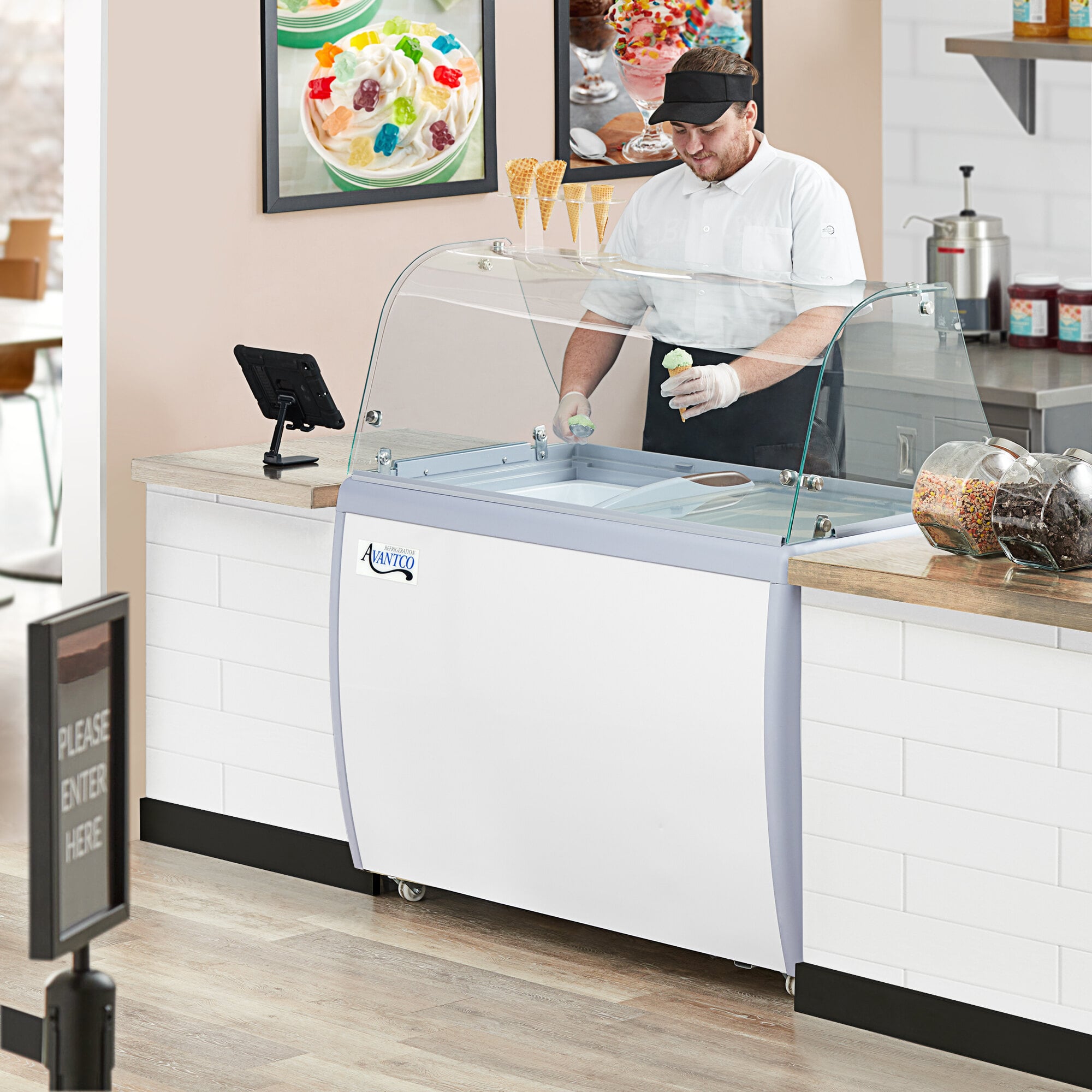 An Avantco ice cream dipping cabinet with a curved sneeze guard on a hospital cafeteria counter.