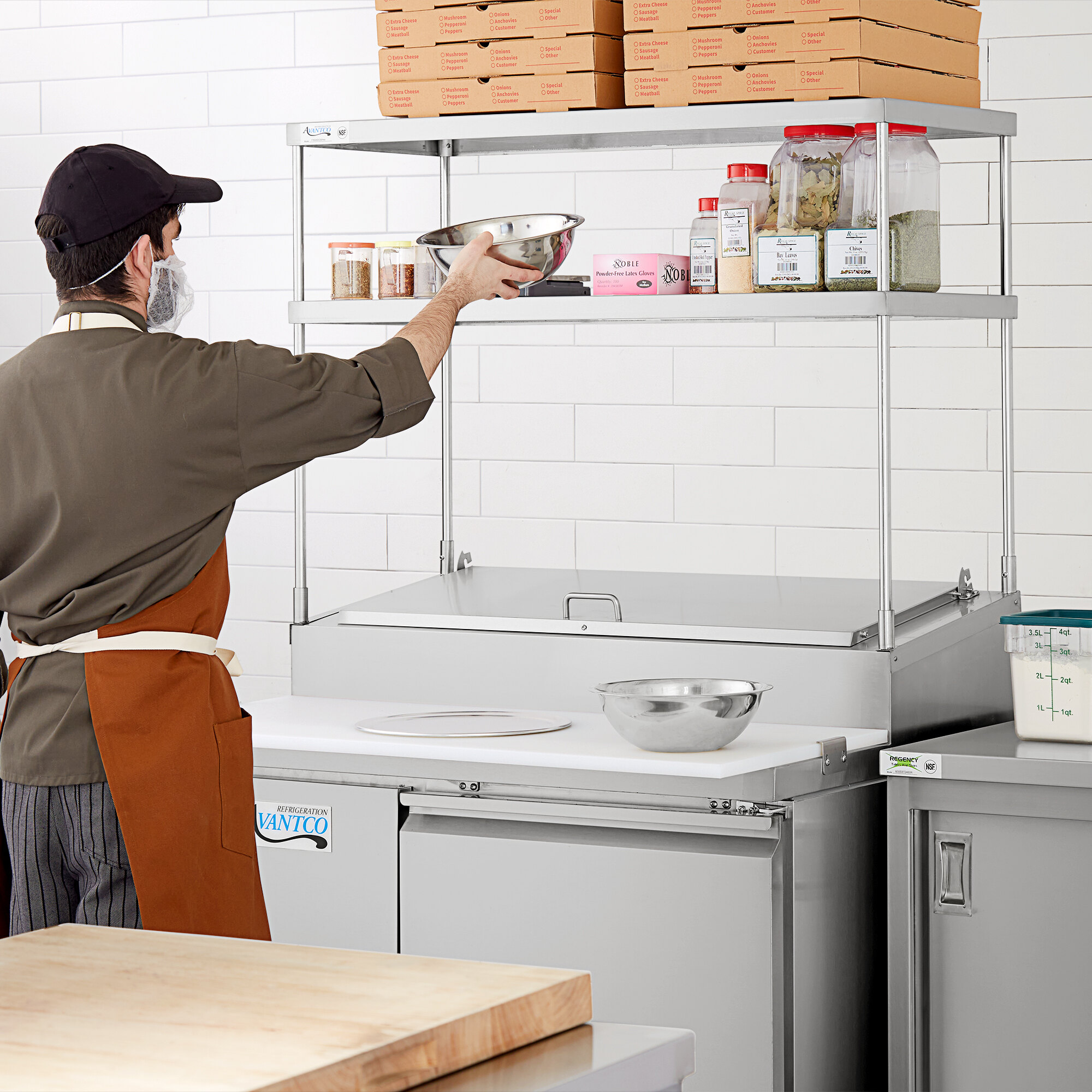 A man in a face mask and apron using a stainless steel double deck overshelf to hold a bowl of food.