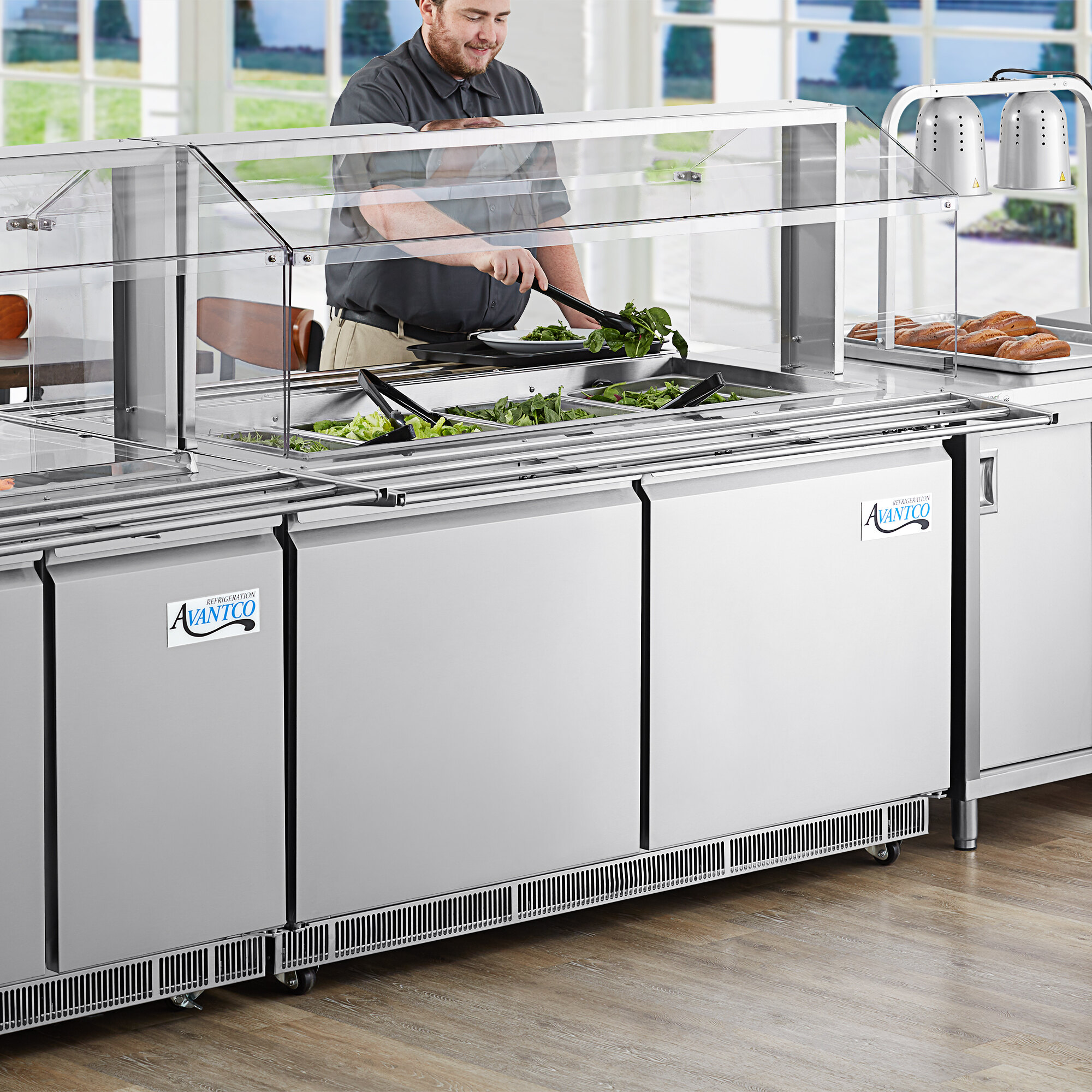 A man standing behind an Avantco Sneeze Guard on a counter in a commercial kitchen.